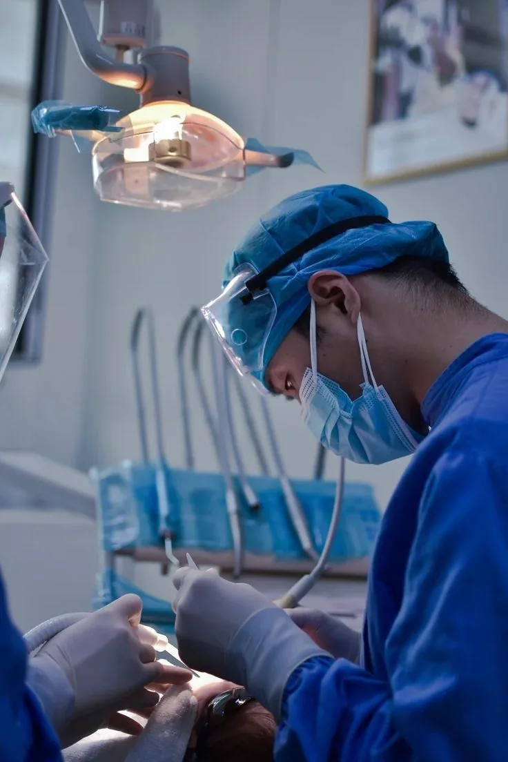 Dentista de touca e máscara azul trabalhando sob o refletor, com óculos de proteção