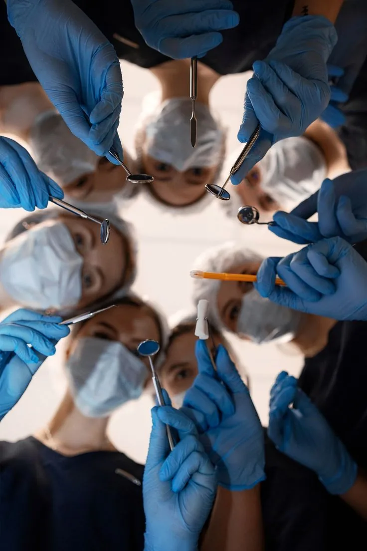 Vista de quem está deitado na cadeira: cinco dentistas de máscara e luvas azuis olhando para baixo, segurando espelhos e instrumentos